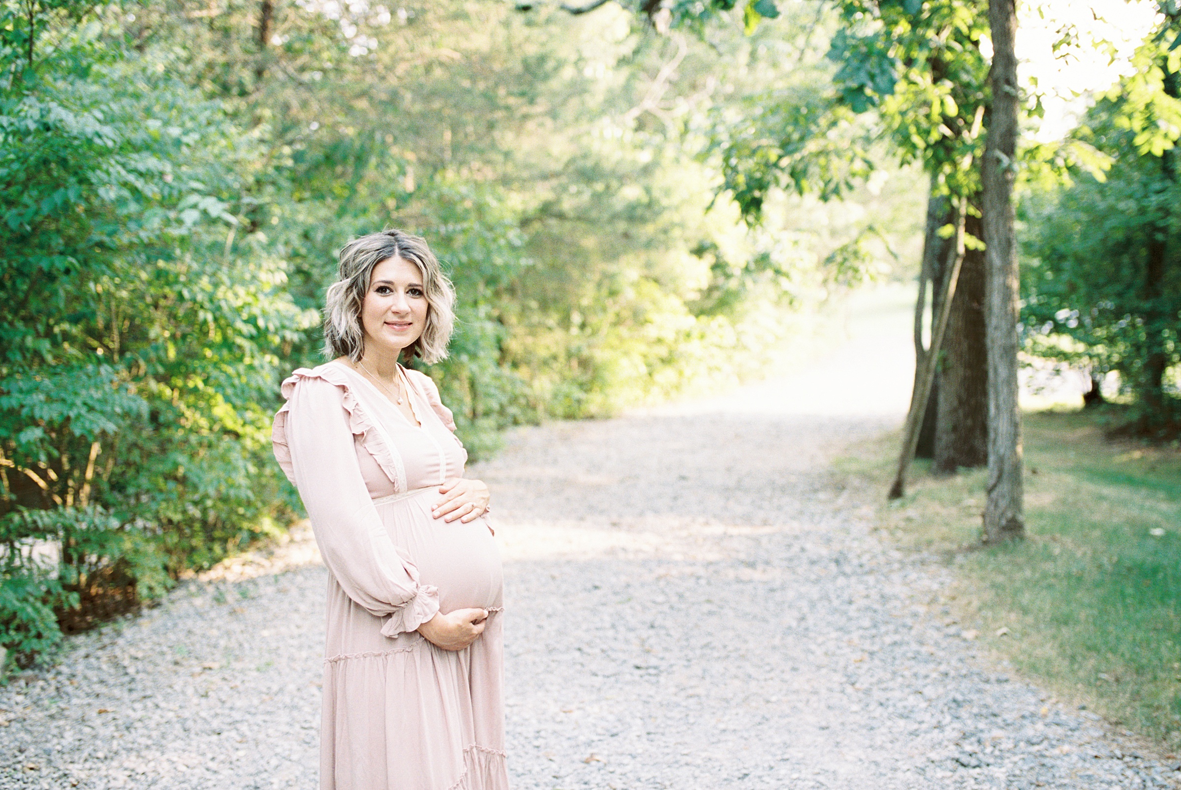 mom-to-be in dusty pink dress holds baby belly on gravel path in Nashville TN