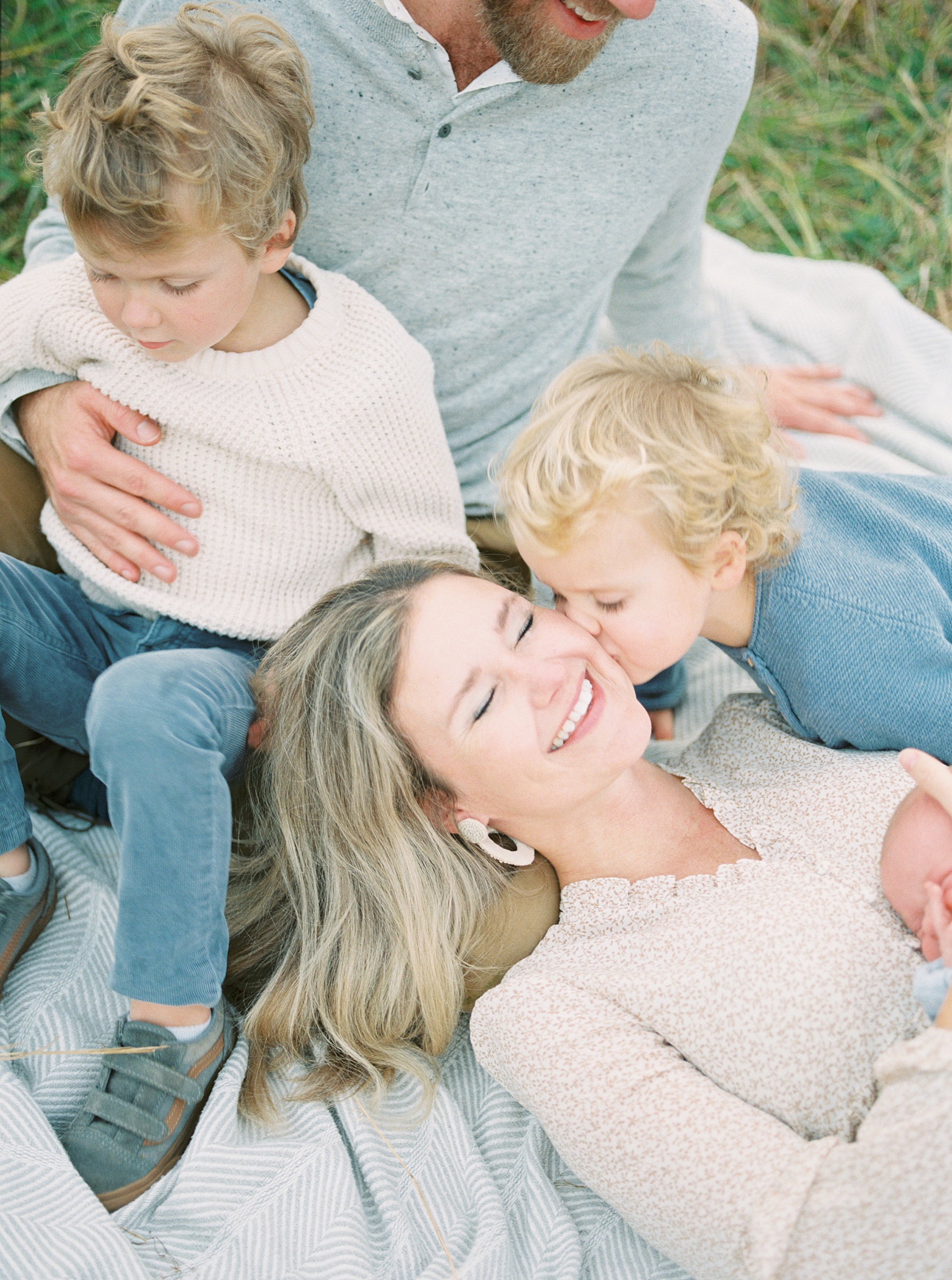 family of five lays in the grass as one of the sons kisses his mom's cheek